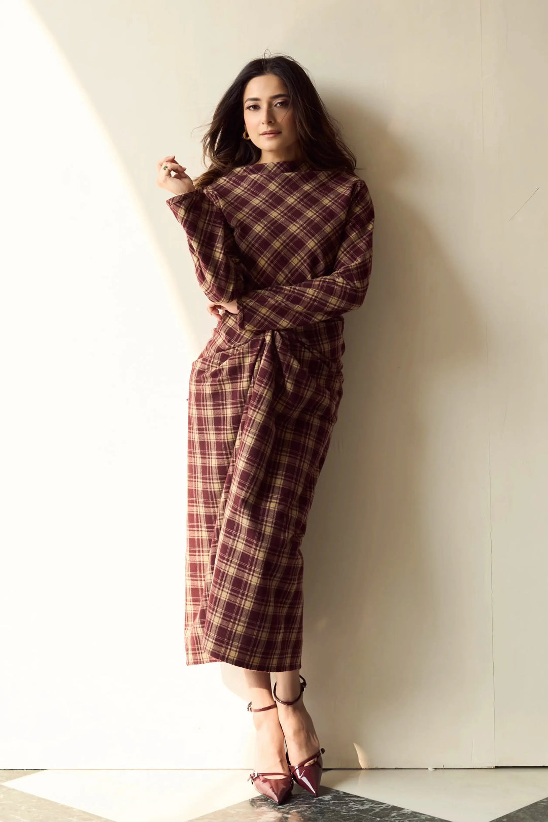 A woman wearing a long-sleeved, high-neck plaid dress in a dark red and brown tartan pattern, leaning against a cream-colored wall.
