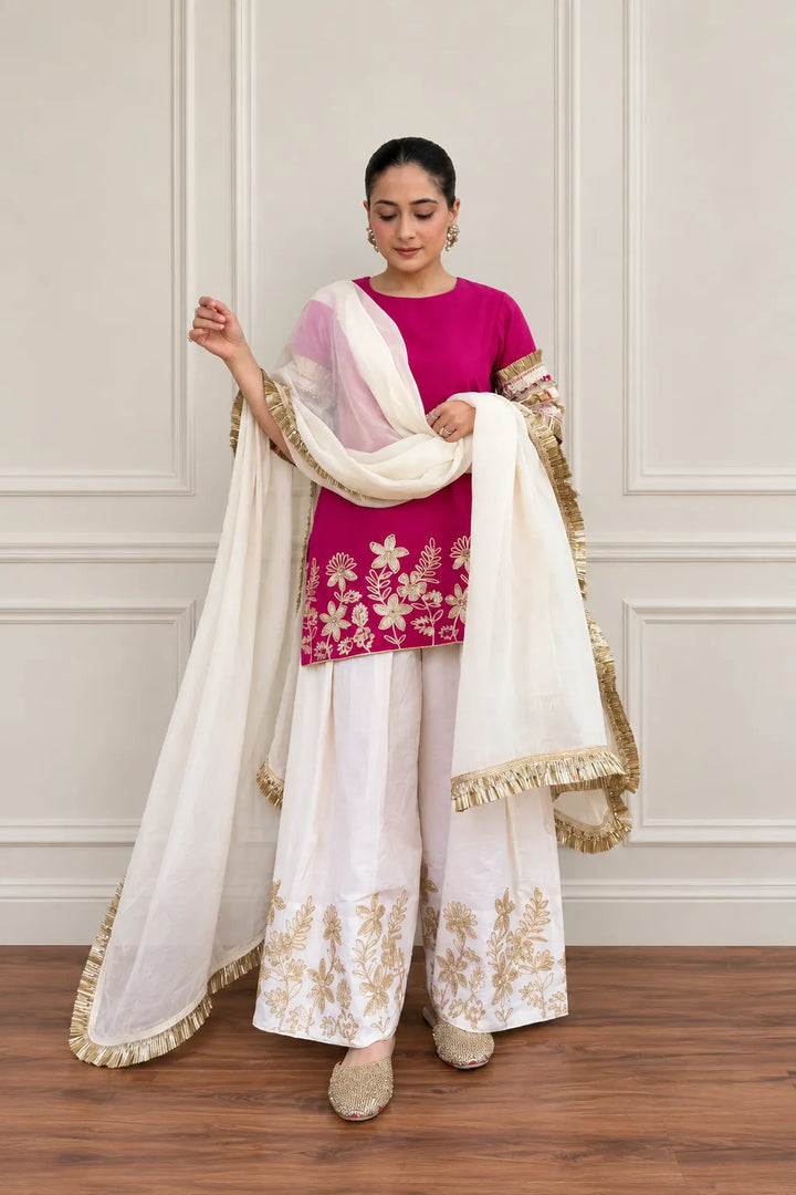 A woman models a traditional Indian outfit consisting of a magenta pink kurta with gold floral embroidery, white flared pants with gold embroidery, and a white dupatta with gold border detailing, set against a neutral paneled wall and wooden floor.