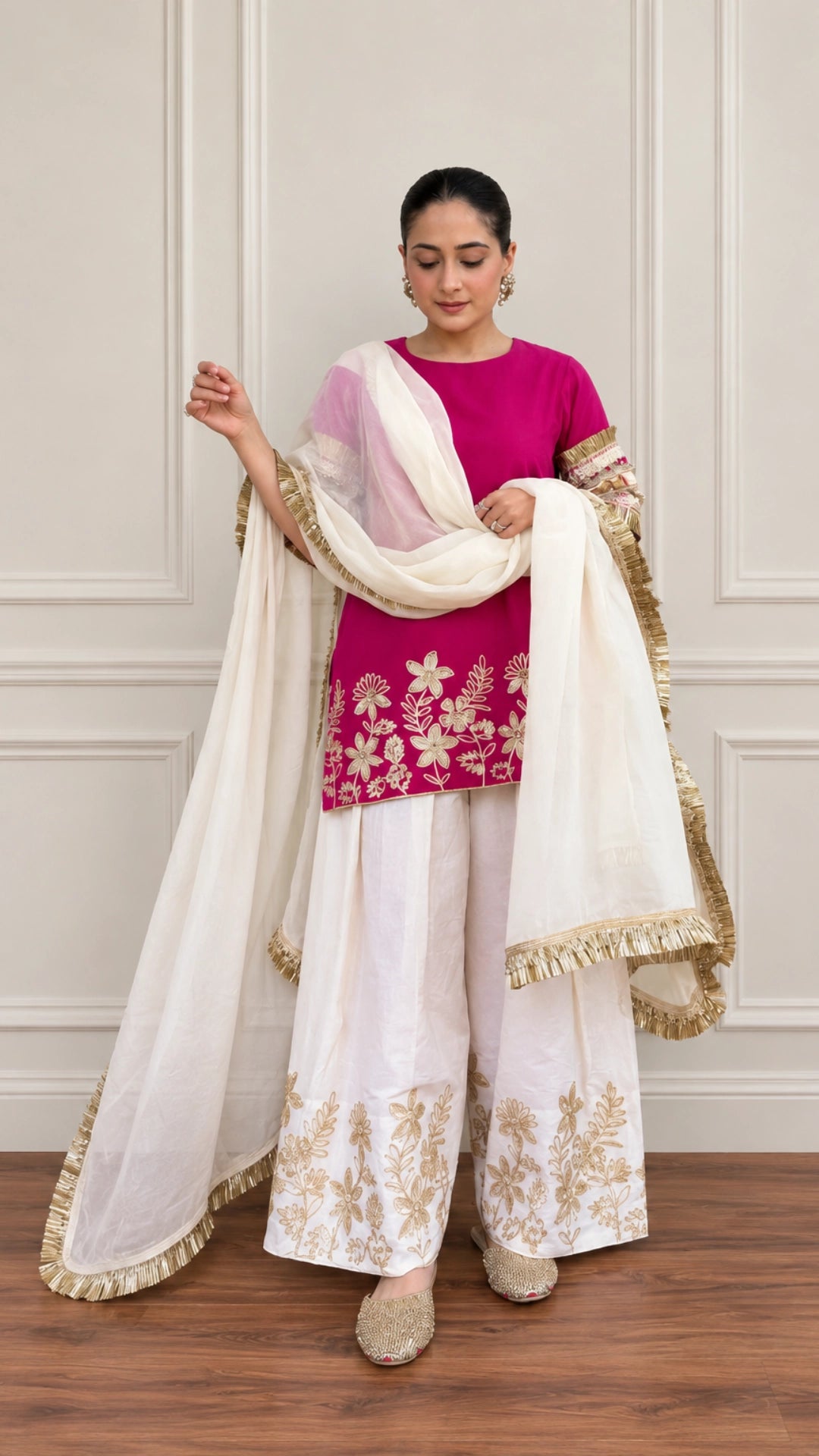 A woman models a traditional Indian salwar kameez suit set. The outfit features a vibrant magenta-pink kameez top with gold floral embroidery, white wide-leg pants with matching embroidery, and a dual-colored white and light pink dupatta with gold borders.