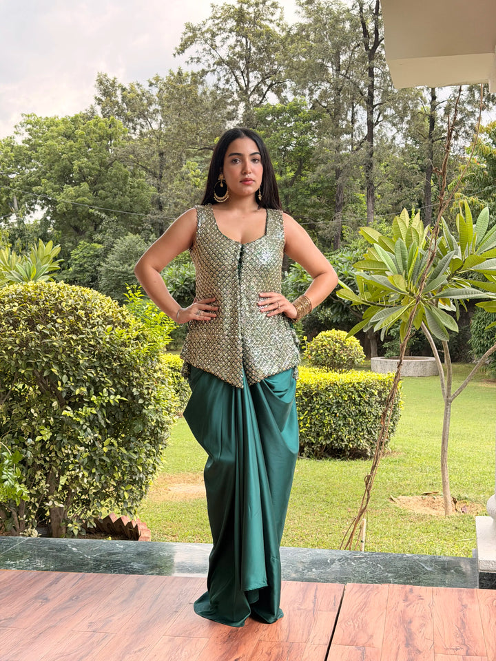 A woman stands outdoors on a wooden deck wearing a sleeveless metallic gold patterned vest with a deep V-neck and a dark green draped skirt, set against a background of green trees and shrubs.