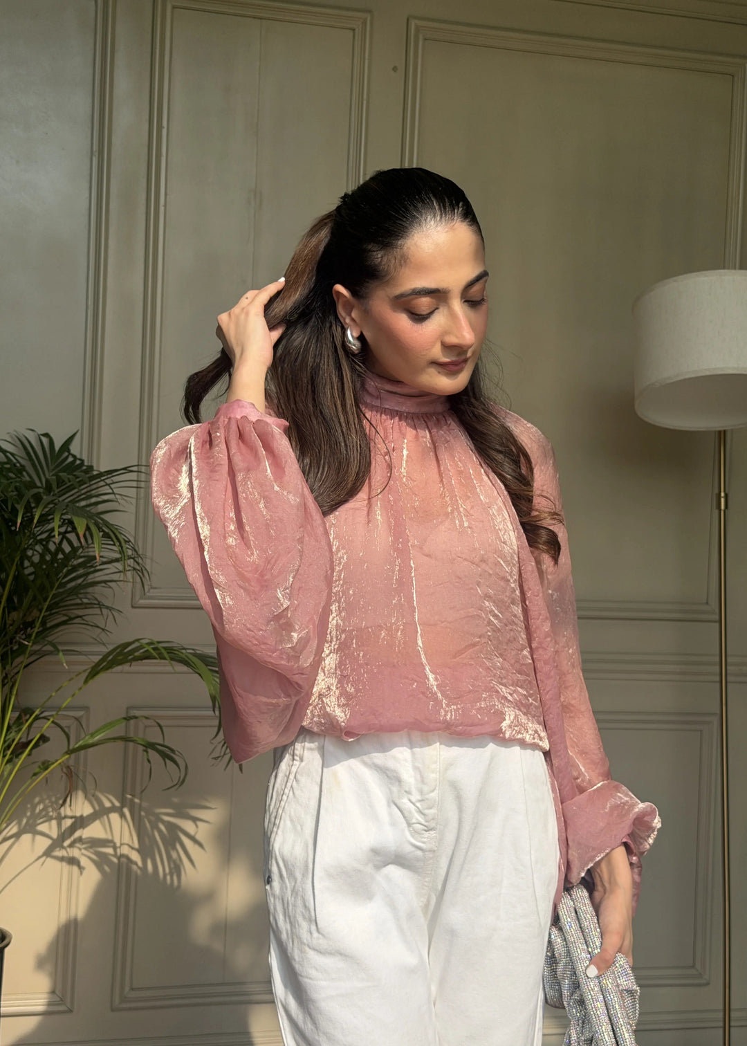A woman wearing a pink textured long-sleeve top and white trousers, standing in an indoor setting with a paneled wall, a plant, and a lamp visible.