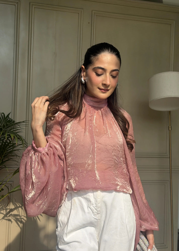 A woman wearing a shiny metallic pink blouse with voluminous sleeves and white trousers, posed in front of a paneled beige wall.