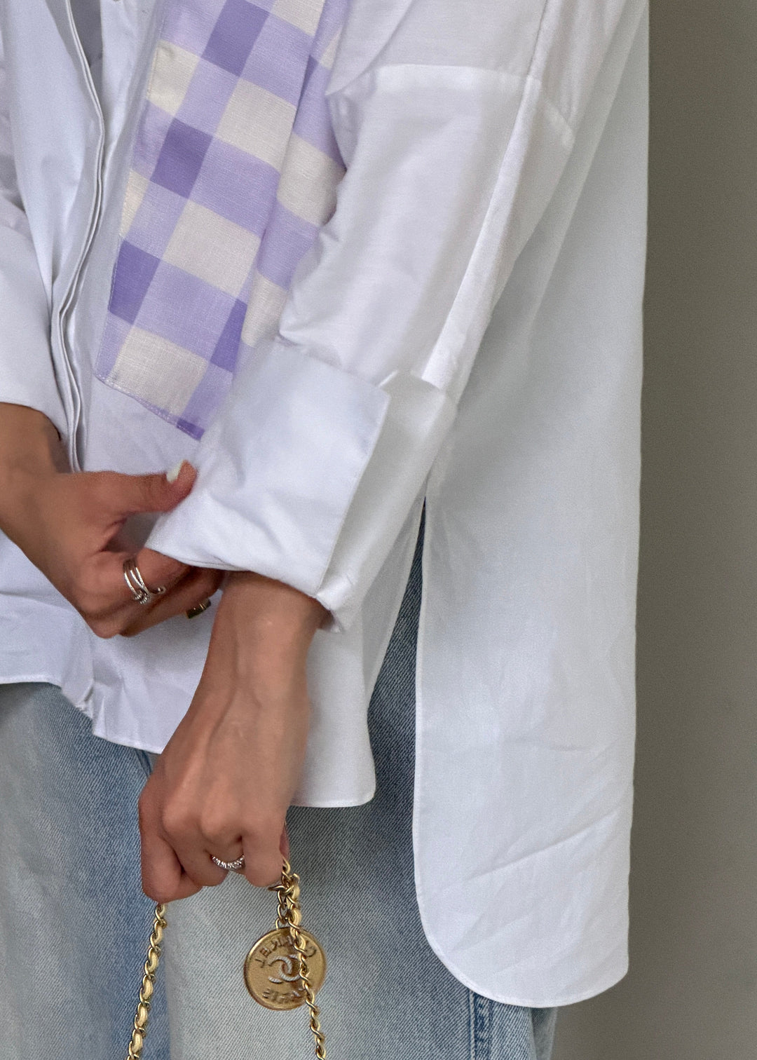 A close-up of a person wearing a white button-up shirt with a lavender and cream gingham patterned chest pocket, paired with light blue jeans and holding a gold chain bag.