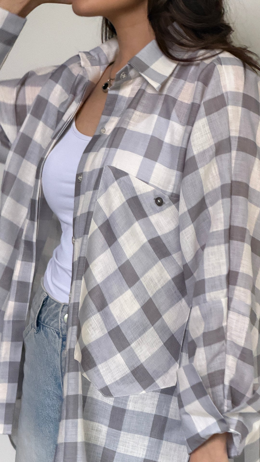 A woman wearing a gray and white plaid button-up shirt over a white tank top and light blue jeans.