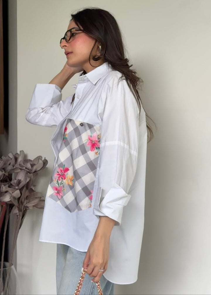 A woman wearing a white oversized button-up shirt with a gray and white gingham patch pocket featuring a pink floral print, paired with light blue jeans and holding a gold chain bag.