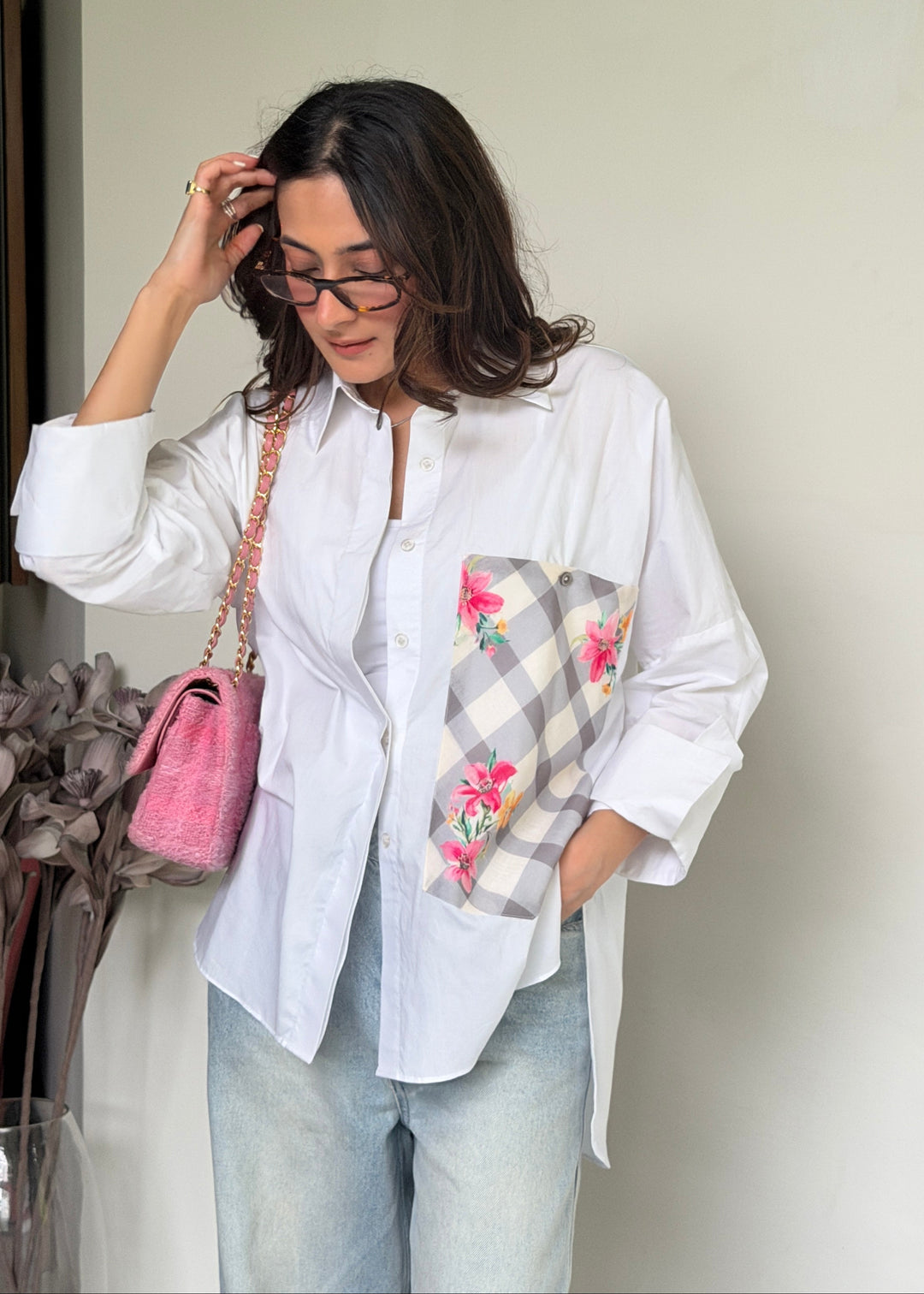 A woman wearing a white button-up shirt with a gray and white plaid pocket featuring pink floral embroidery, paired with light blue jeans and a pink handbag.
