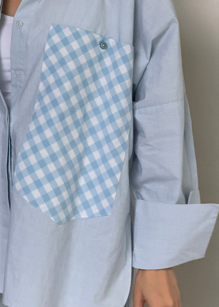 A light blue oversized button-up shirt with a large light blue and white gingham pocket on the chest, worn by a model.