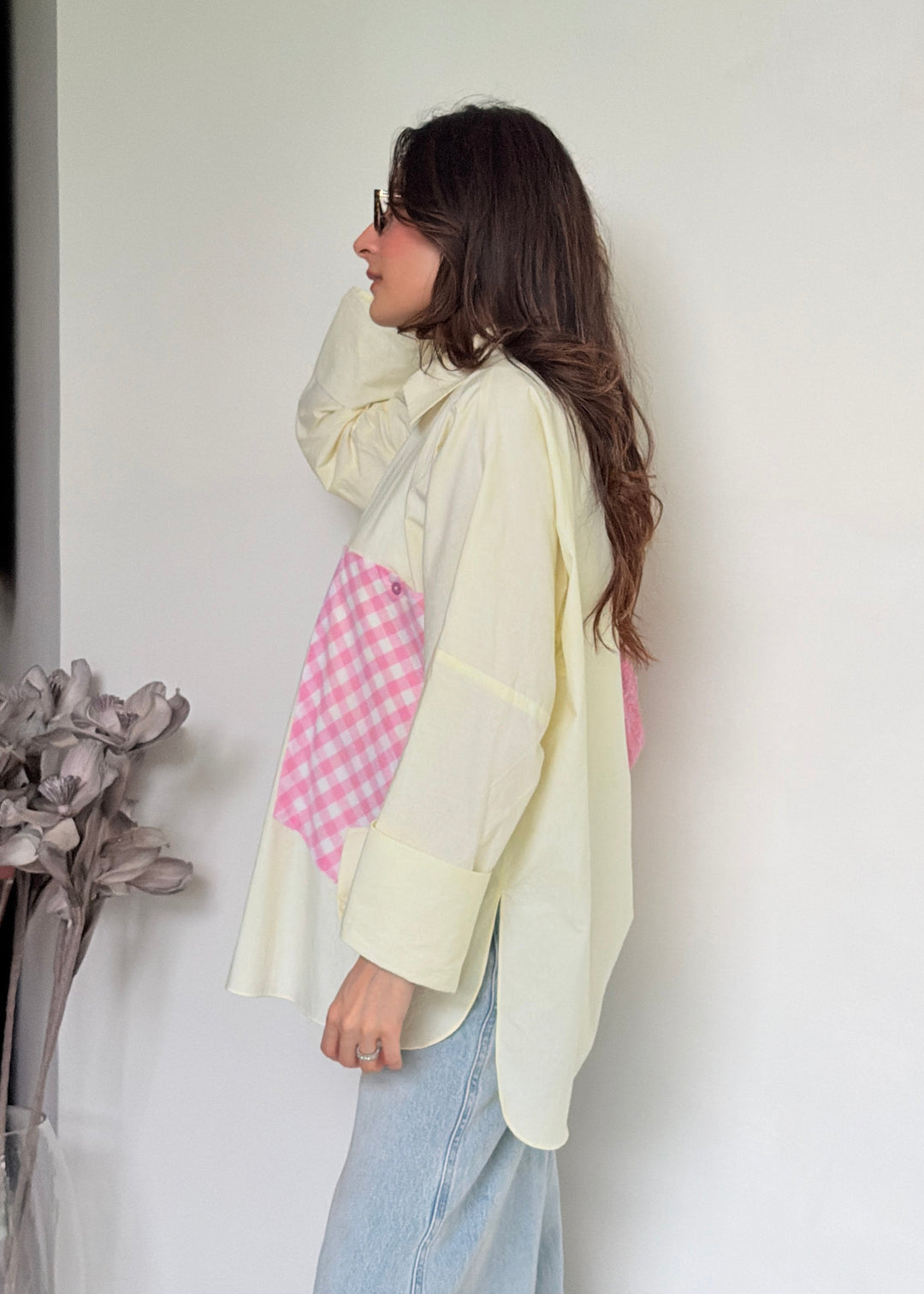 A woman wearing a cream-colored patchwork shirt with pink gingham panels, paired with light blue jeans, standing against a plain white wall.