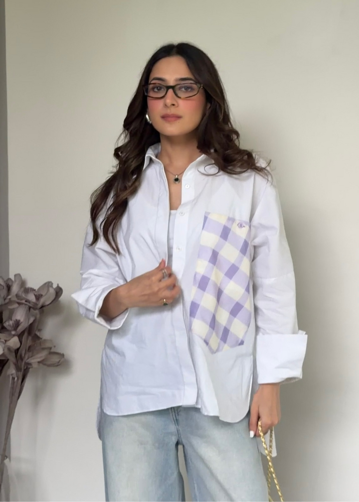 A woman wearing a white button-up shirt with a purple and white gingham pocket detail, paired with light blue jeans, standing against a plain off-white wall.