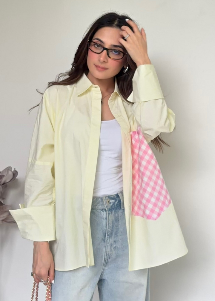 A woman wearing a pale yellow button-up shirt with a pink and white gingham patch pocket, layered over a white tank top and light blue jeans, standing against a plain off-white wall.