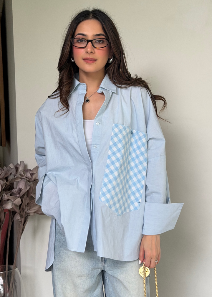 A woman wearing a light blue button-up shirt with a light blue and white gingham pocket on the chest and a matching gingham pattern on the right sleeve, paired with light-wash jeans.