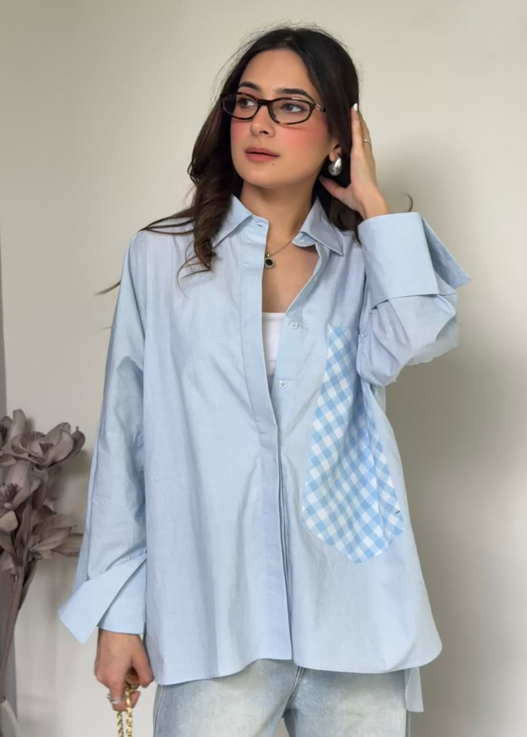 A woman wearing a light blue button-up shirt with a blue and white gingham pocket, paired with light-wash jeans and glasses, standing against a plain beige wall.