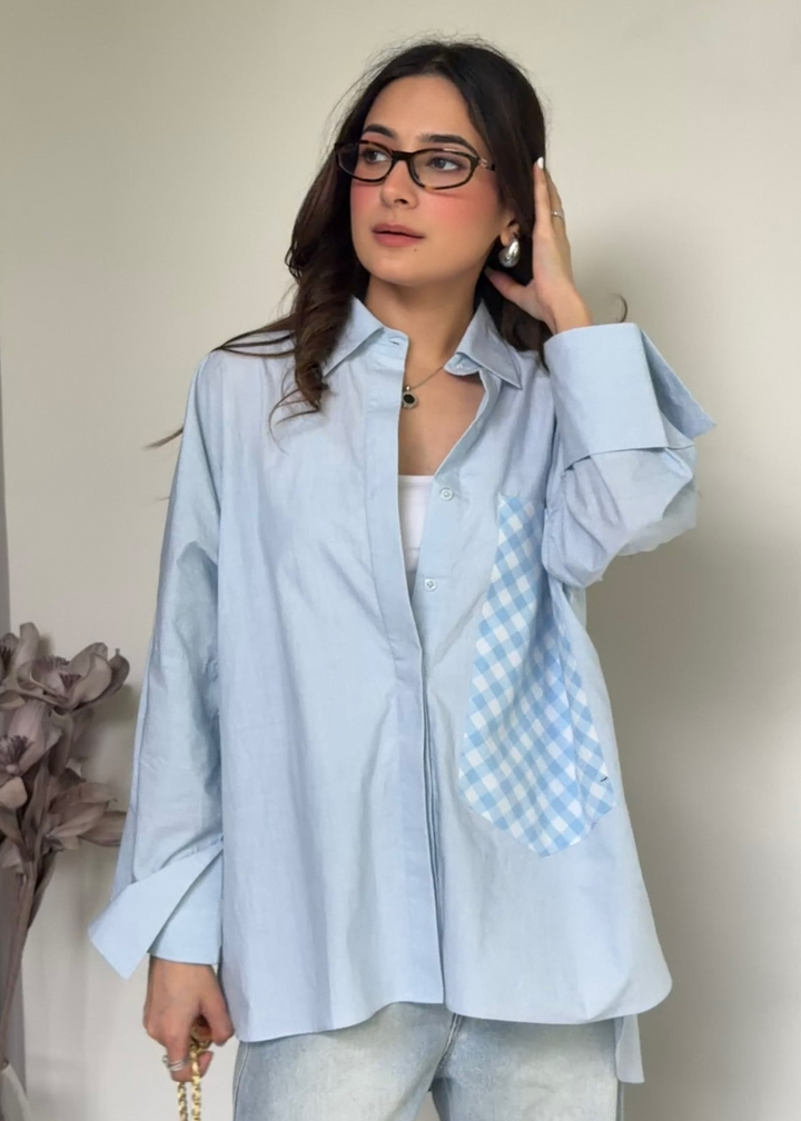 A woman wearing a light blue button-up shirt with a blue and white gingham pocket, paired with light-wash jeans and glasses, standing against a plain beige wall.