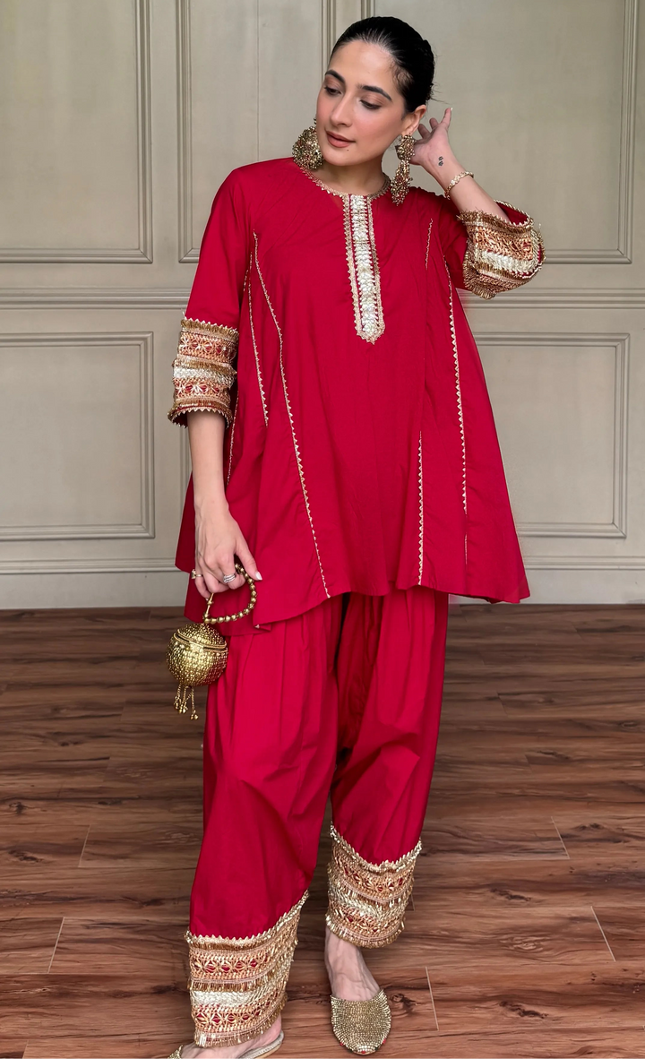 A woman modeling a bright red Indian ethnic suit with gold embroidery, featuring a kurta, pants, and a small gold handbag, set against a paneled beige wall and wooden floor.