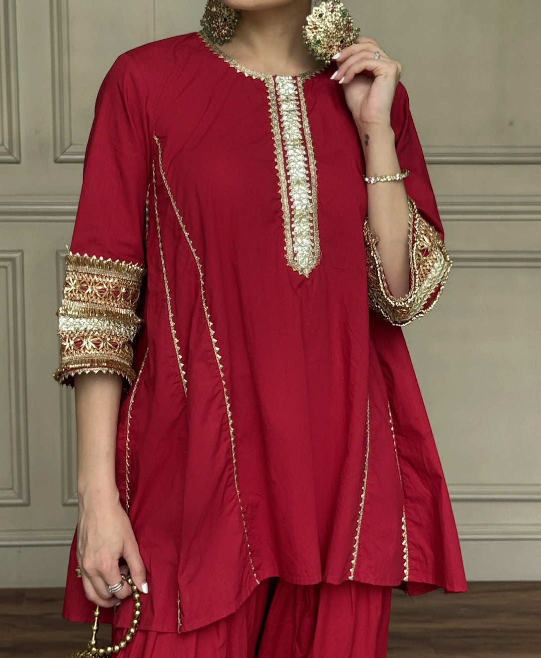 A woman wearing a deep red kurta with ornate gold embroidery on the neckline, sleeve cuffs, and side seams, paired with matching red trousers. She is holding a beaded necklace and standing against a neutral paneled wall.