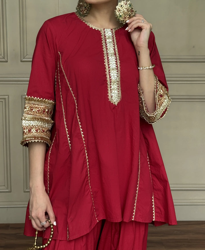 A woman wearing a deep red kurta with ornate gold embroidery on the neckline, sleeve cuffs, and side seams, paired with matching red trousers. She is holding a beaded necklace and standing against a neutral paneled wall.