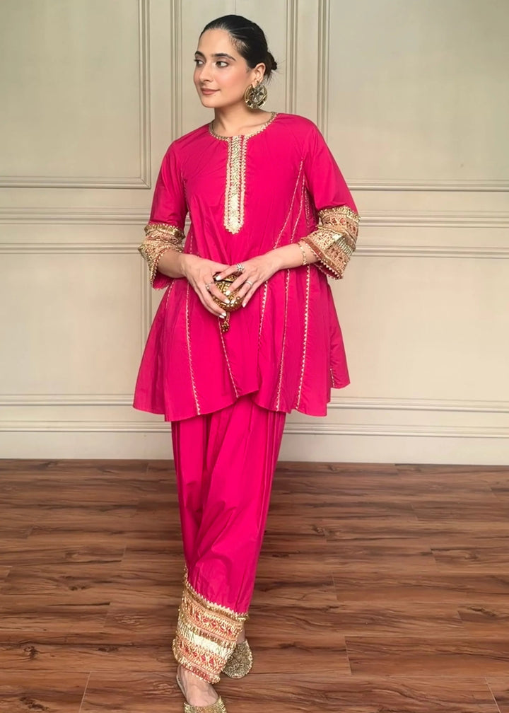 A woman modeling a bright pink traditional South Asian outfit with gold embroidery, standing against a beige paneled wall and wooden floor.