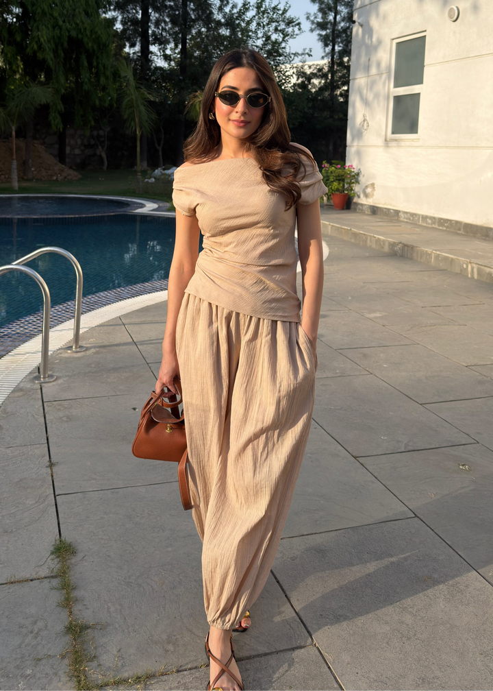 Woman in a beige outfit standing by a poolside with trees and a building in the background.