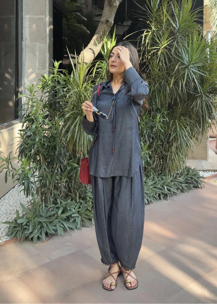 Woman in a gray outfit standing outdoors with plants and a building in the background