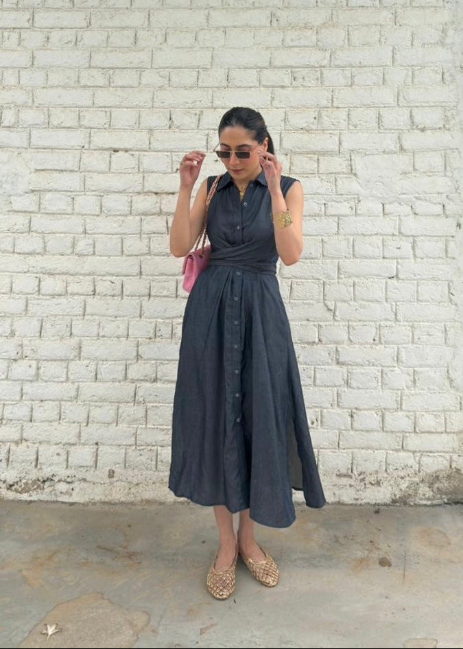 A woman wearing a dark gray sleeveless midi dress with a button-down front and a matching belt, standing against a white painted brick wall.