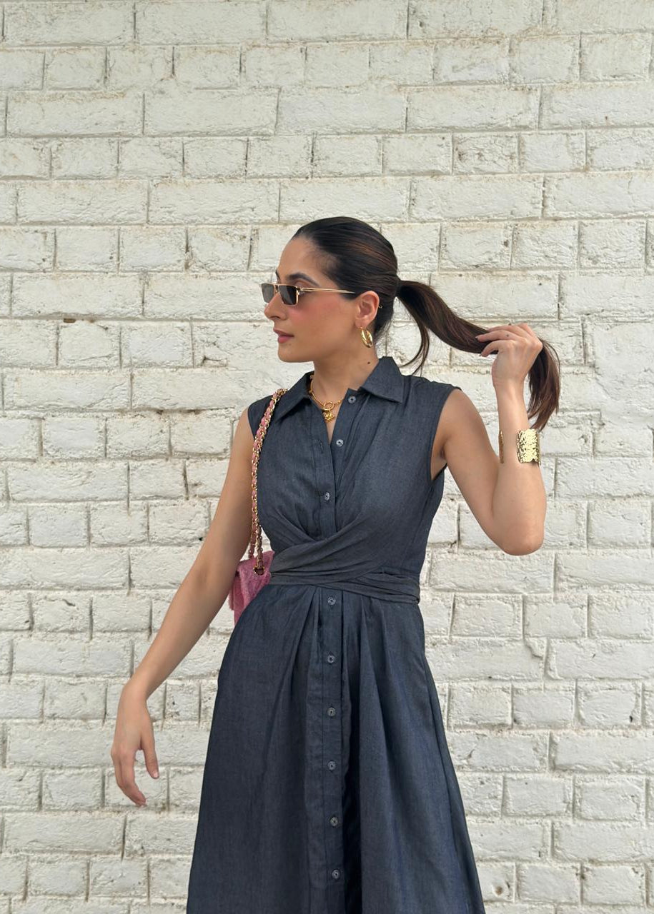 A woman wearing a sleeveless dark slate blue button-down midi dress with a wrap-style waist, standing against a white painted brick wall.