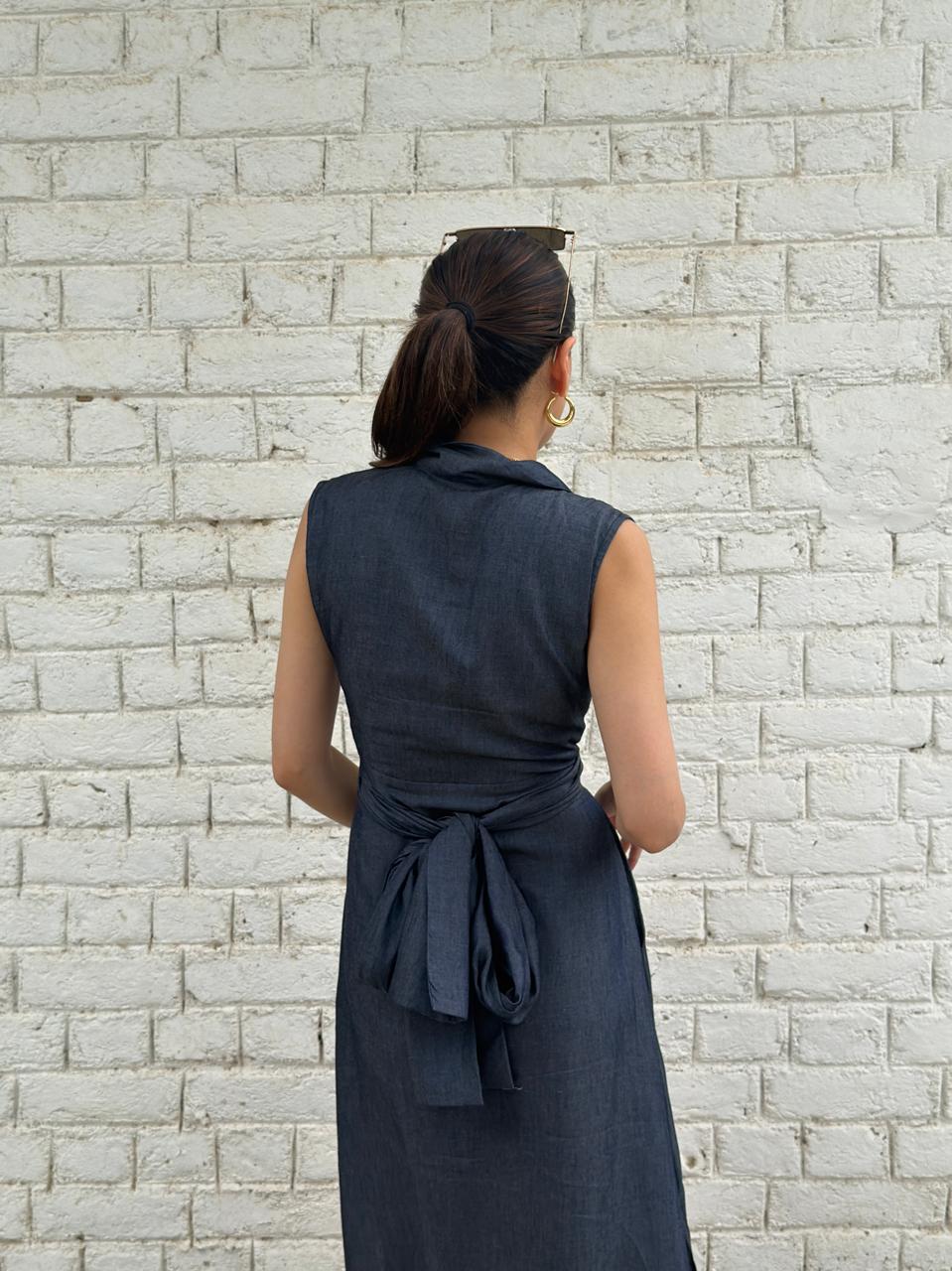 A woman viewed from behind wearing a sleeveless dark navy wrap-style dress with a tied waist, standing against a white painted brick wall.