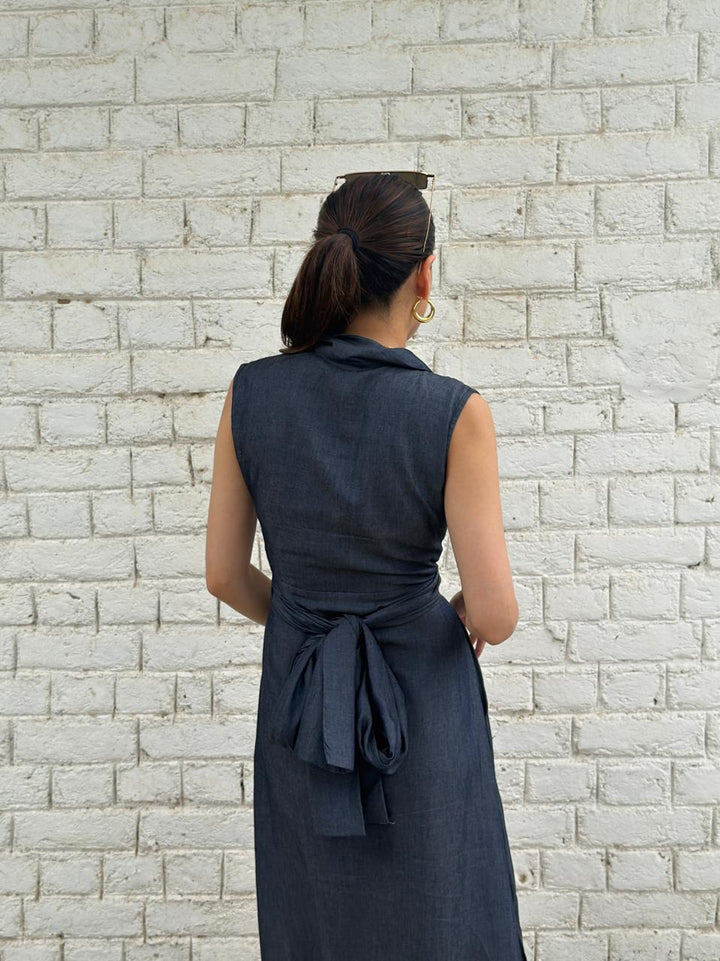 A woman viewed from behind wearing a sleeveless dark navy wrap-style dress with a tied waist, standing against a white painted brick wall.