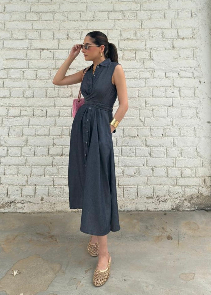 A woman wearing a sleeveless dark gray midi dress with a tie waist, standing in front of a white brick wall.