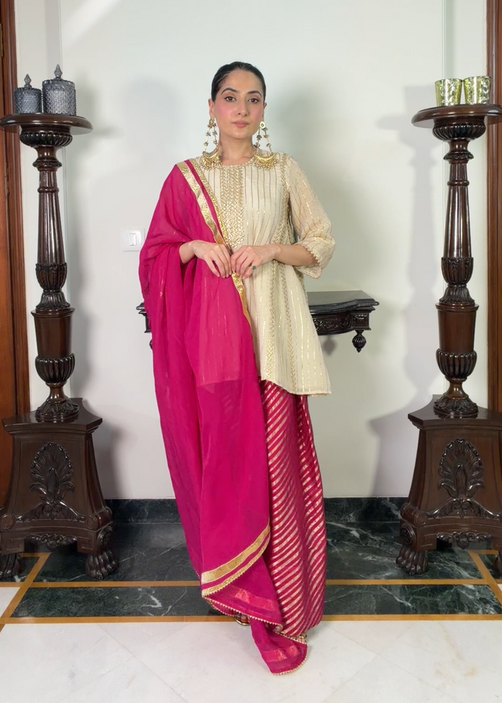 A woman modeling a traditional Indian outfit consisting of a beige kurta, a bright pink dupatta, and a pink skirt with gold stripes, standing in an indoor room with decorative pillars.