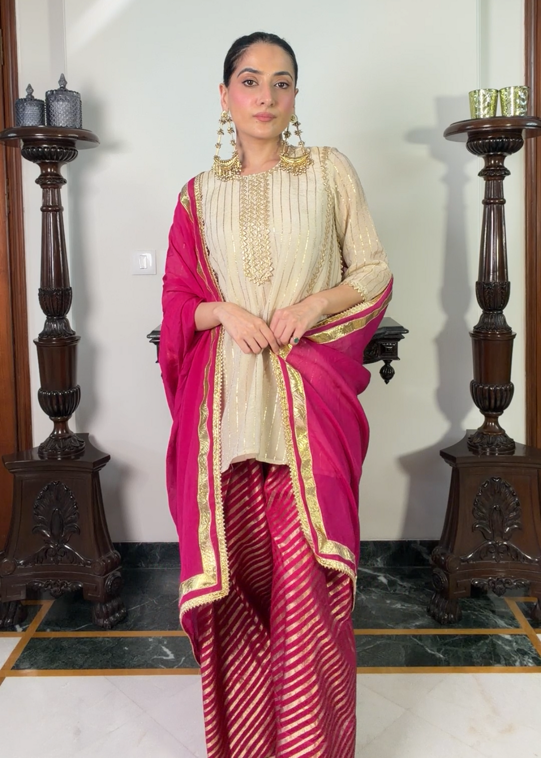 A woman modeling a traditional Indian outfit consisting of a beige embroidered kurta, a bright pink dupatta, and a pink and gold patterned saree, standing in an indoor setting with dark wooden pillars.