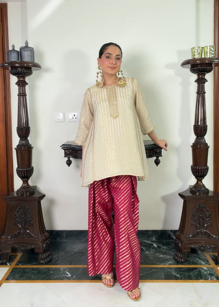 A woman modeling a cream-colored embroidered kurta paired with magenta and white striped palazzo pants, standing in an indoor setting between two dark wooden pillars.