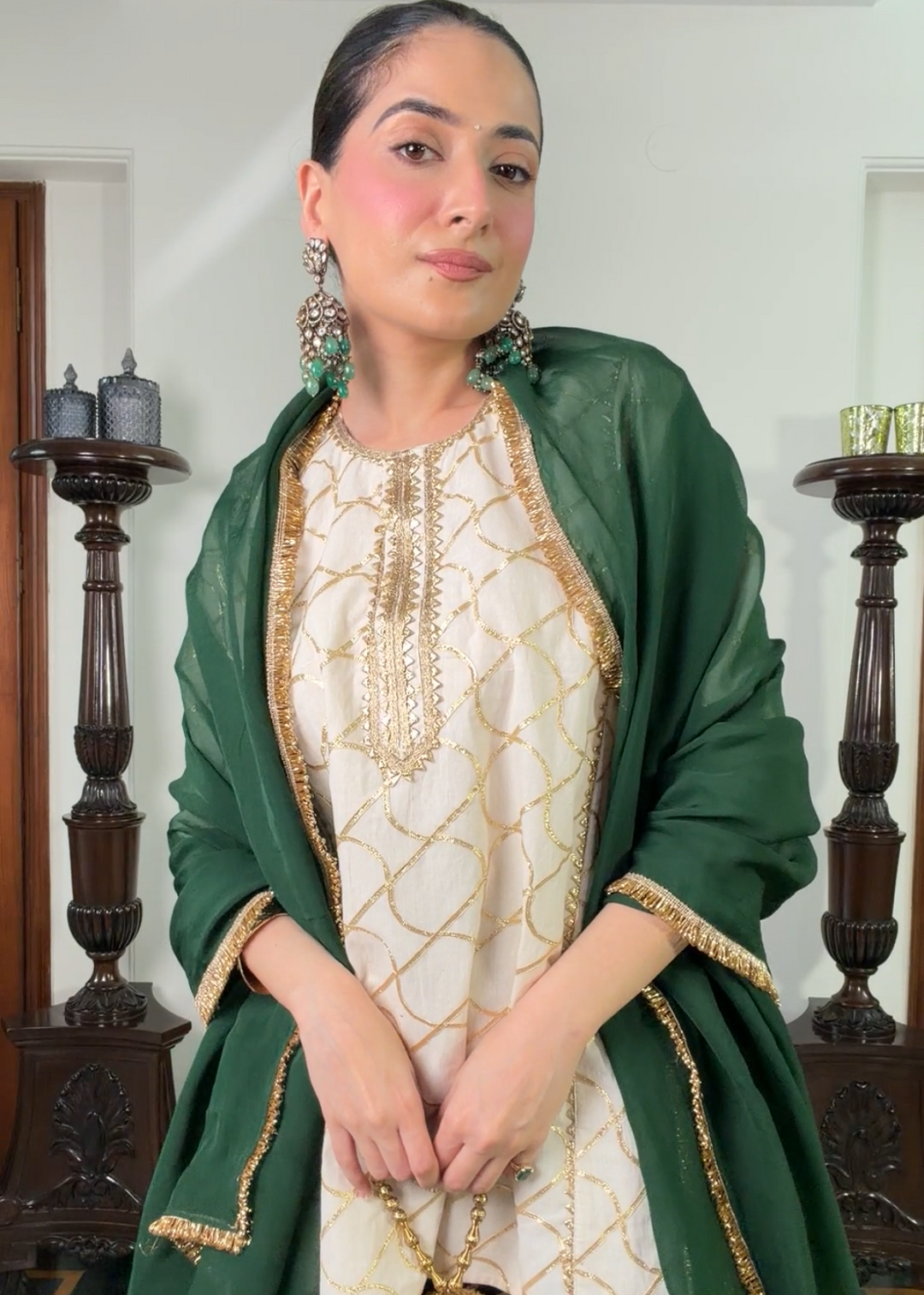 A woman modeling a traditional South Asian outfit featuring a cream-colored embroidered kurta with gold detailing, a green dupatta with gold borders, and green striped trousers, set against an indoor background with wooden pillars.