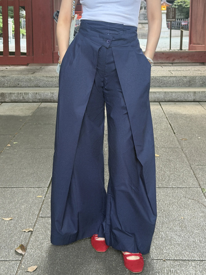 A person wearing high-waisted, wide-leg navy blue pants with a wrap-style front detail, paired with a white top and red shoes, standing outdoors on stone pavement.