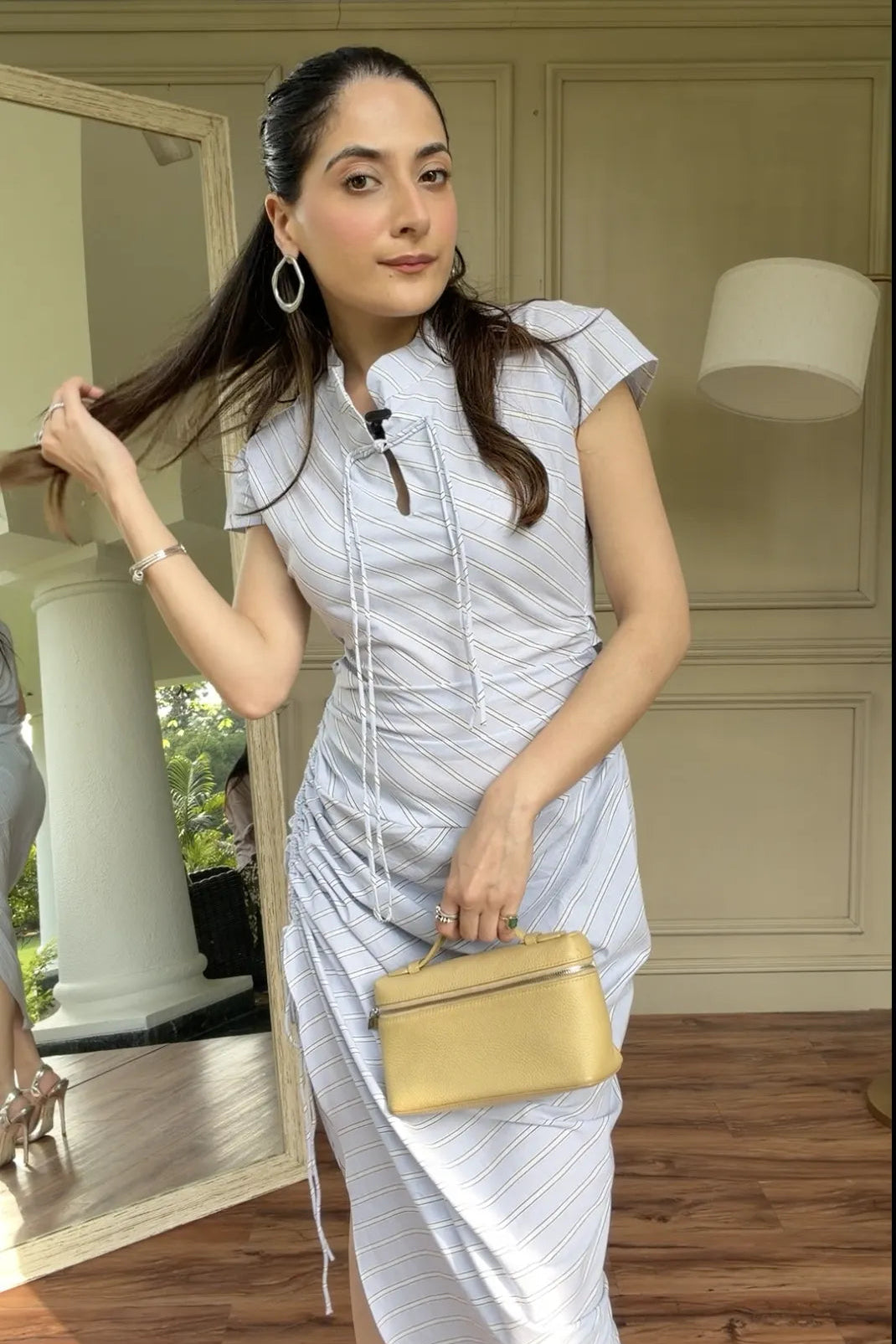 A woman models a light blue and white vertically striped midi dress with a high collar, short sleeves, and a drawstring tie at the neckline. She is holding a small gold handbag in an indoor setting with a wooden floor and a mirror.