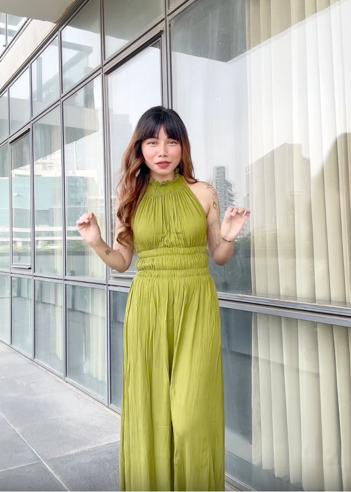 A woman wearing a sleeveless olive green halter maxi dress stands in front of a modern glass building facade.