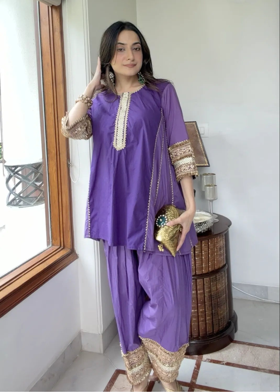 A woman modeling a purple traditional outfit with gold embroidery, standing in a bright indoor room near a window.