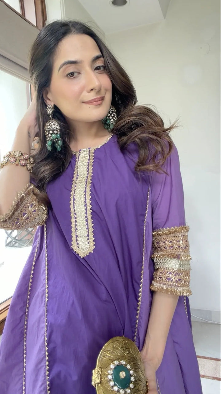 A woman wearing a purple traditional outfit with ornate gold embroidery on the neckline and sleeve cuffs, holding a gold clutch with a green stone, in an indoor setting.