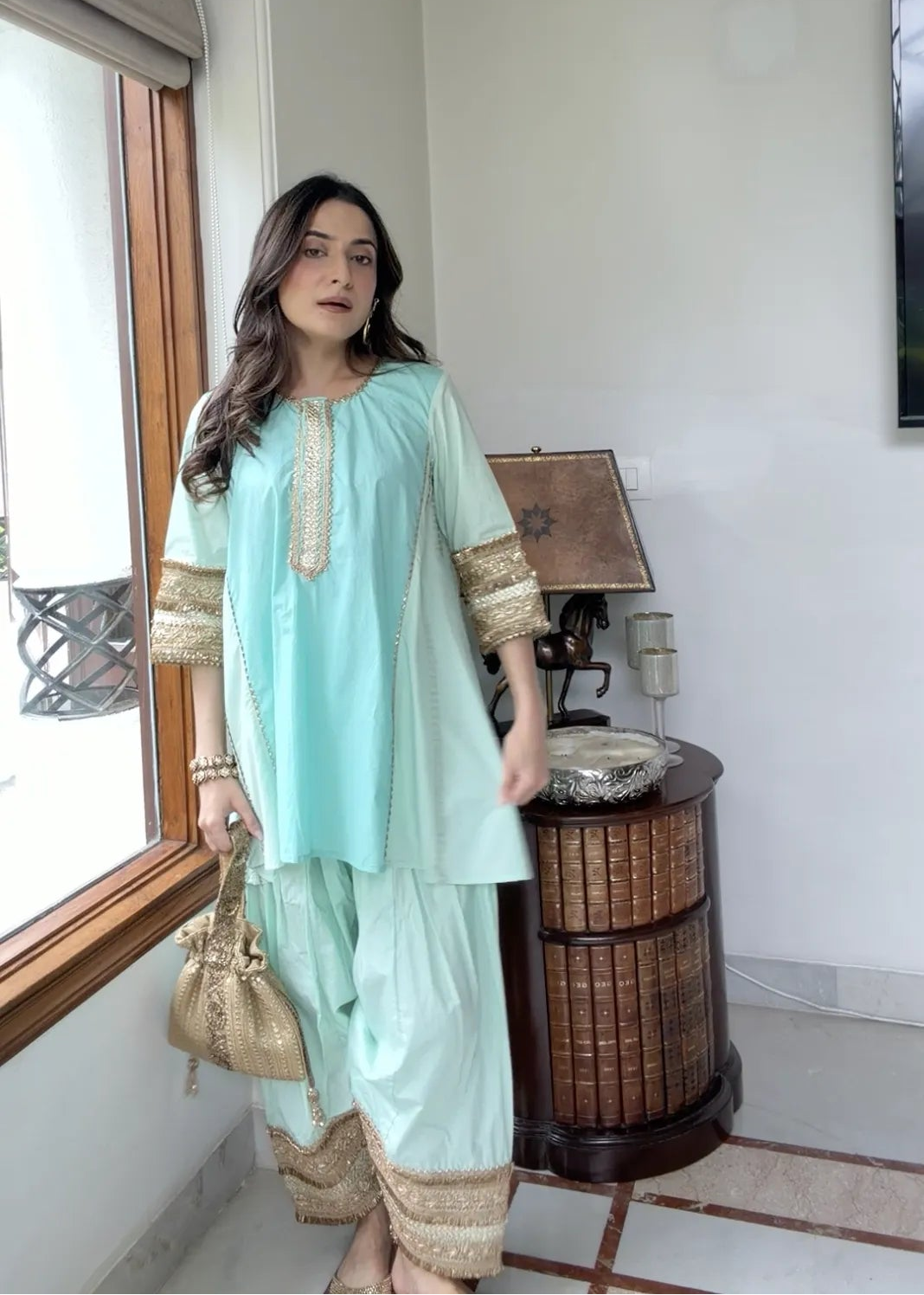 A woman models a mint green traditional three-piece salwar kameez set with gold embroidery, standing in a brightly lit room next to a window.