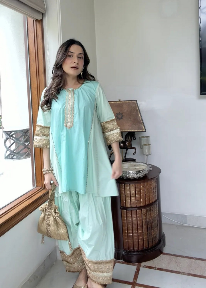 A woman models a mint green traditional three-piece salwar kameez set with gold embroidery, standing in a brightly lit room next to a window.