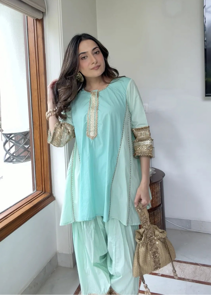 A woman models a mint green traditional South Asian outfit with gold embroidery, paired with matching pants and holding a gold clutch, standing in a brightly lit indoor space.