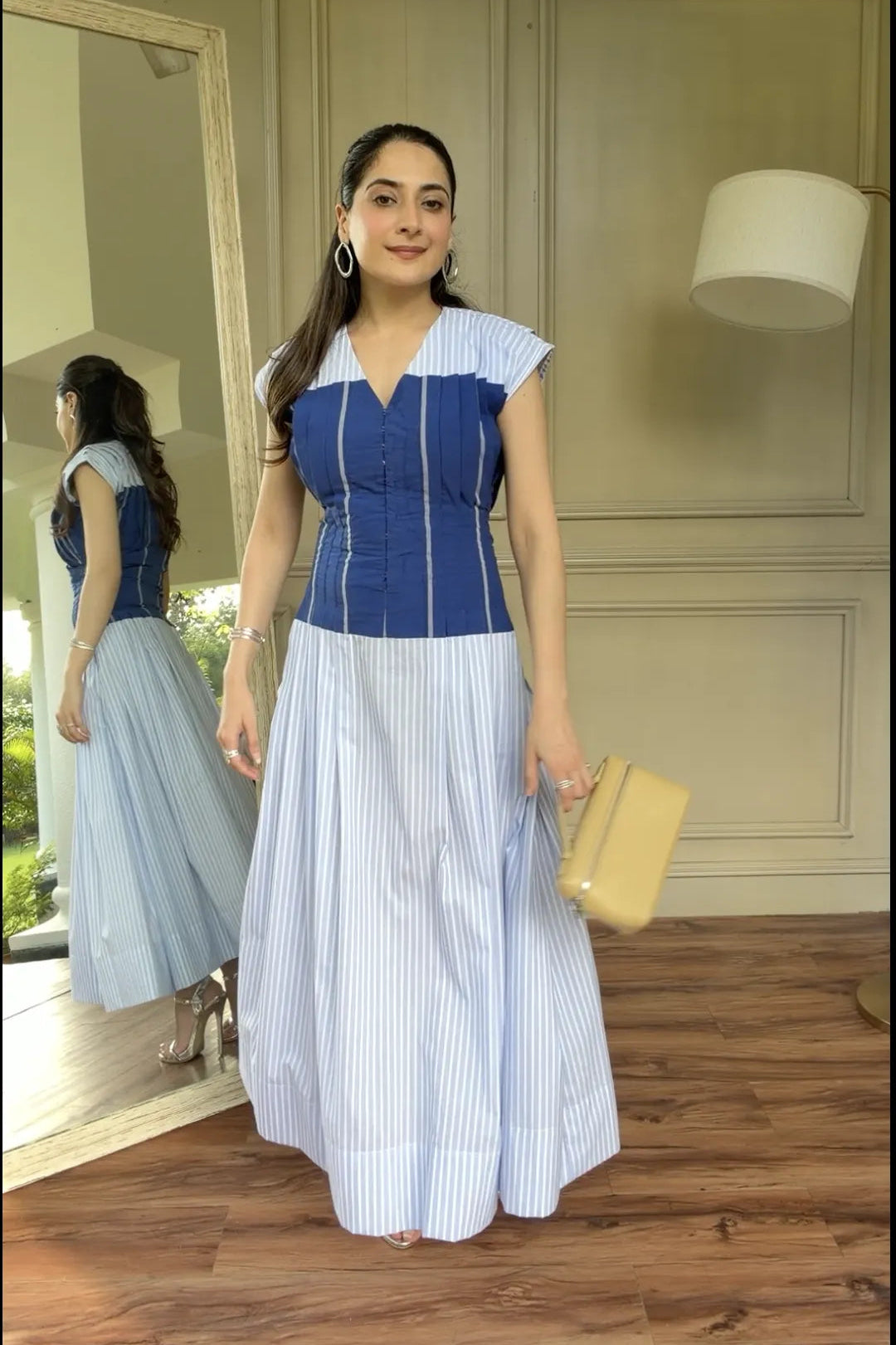 A woman modeling a sleeveless maxi dress with a blue corset-style bodice and a light blue and white vertically striped skirt, standing in an indoor room with wood floors and a mirror showing her back.