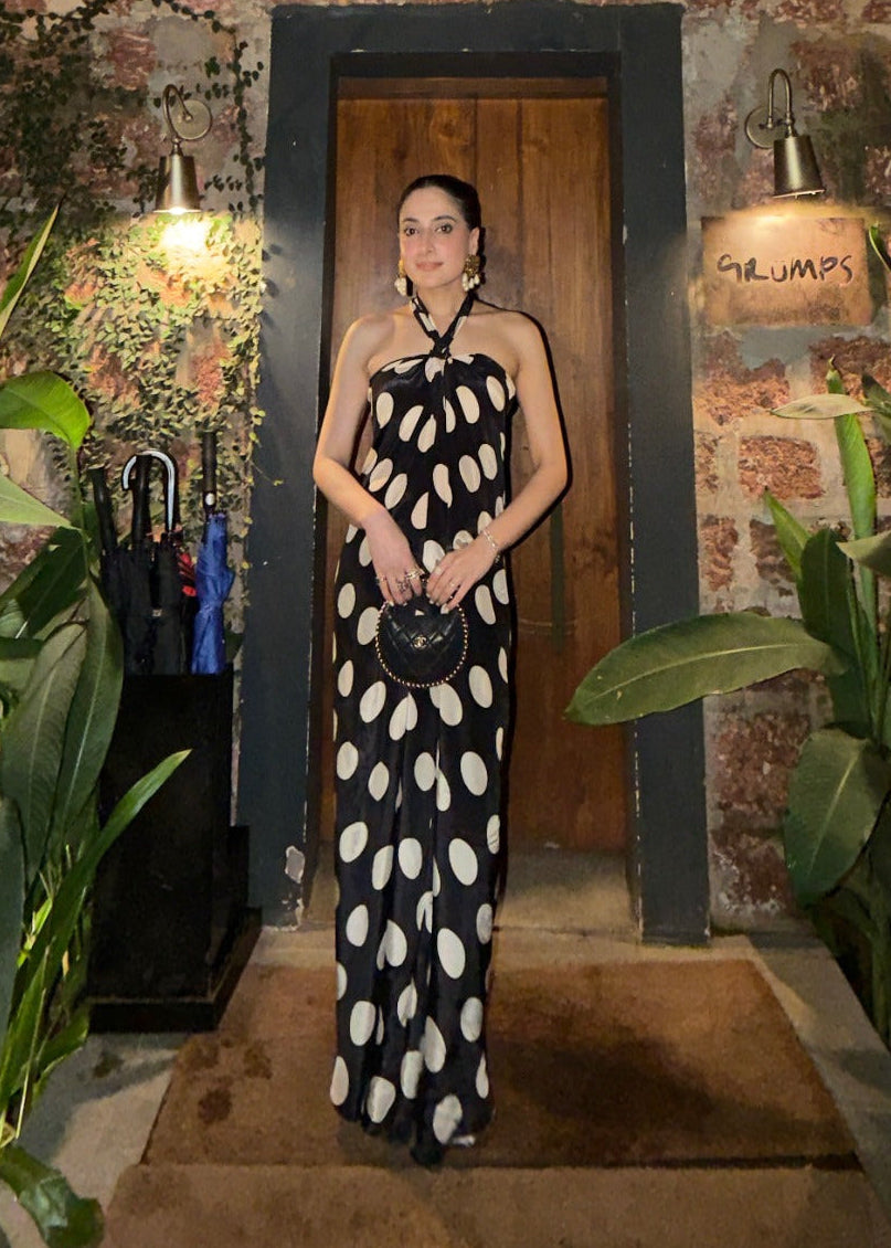 A woman stands in a doorway wearing a black halter-neck maxi dress with large white polka dots. She is holding a small black handbag.